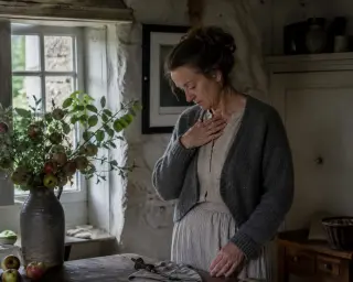 Middle-aged woman in a cozy Normandy kitchen, quietly reflecting by a window, hand resting over her chest, with soft natural morning light and rustic details.