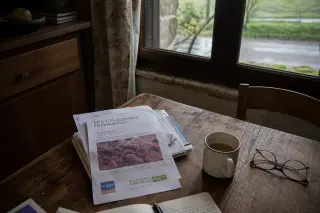 Medical leaflets and reading glasses on a rustic wooden kitchen table with soft morning light and a glimpse of Normandy countryside through the window.