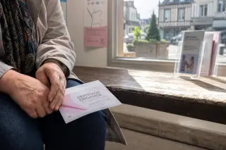 Close-up of a woman's hands holding a medical invitation letter on her lap in a softly lit Normandy health center waiting area, with morning light streaming through a window onto a wooden bench.